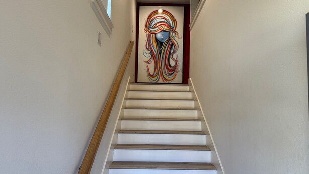 Una escalera con peldaños de madera y paredes blancas, que conduce a una puerta con un cuadro abstracto y colorido.
