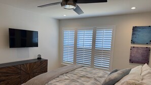 Un dormitorio con cama, televisión, ventilador de techo y ventana con persianas.