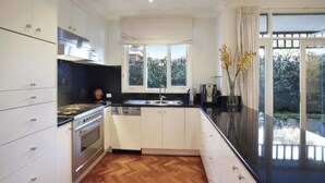 A modern kitchen with white cabinets, a black countertop, and a wooden floor. There is a window providing natural light and a view of greenery outside. - Luxurious Kew House - 5 min walk to cafes and restaurants (Kew)