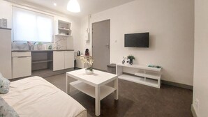 Ein kompakter, moderner Raum mit Bett, kleinem Tisch mit Blumenvase, einer Küchenzeile und einem an der Wand befestigten Fernseher.