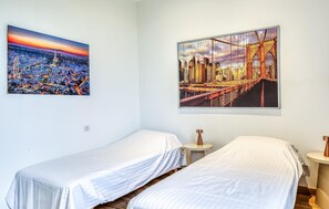 A hotel room with two single beds, a nightstand, and two wall-mounted paintings depicting cityscapes. - Gorgeous home in Auvilliers with WiFi (Auvilliers)