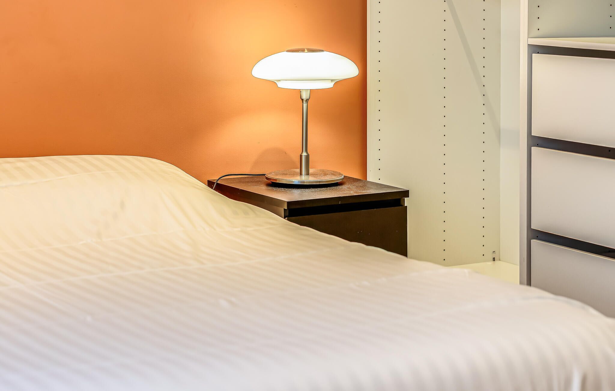 Un lit avec un drap blanc, une table de chevet avec une lampe et un mur avec une étagère.