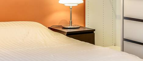 Un lit avec un drap blanc, une table de chevet avec une lampe et un mur avec une étagère.