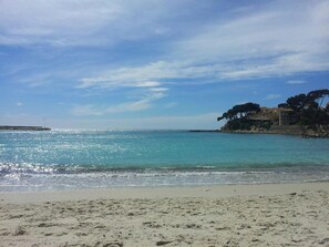 Ein Strand mit klarem türkisblauem Wasser, einem sandigen Ufer und einem Gebäude mit Bäumen auf einer Klippe.