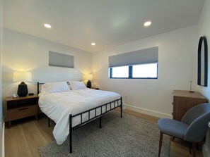 A bedroom with a bed, two bedside tables with lamps, a chair, a dresser, and two windows with blinds.