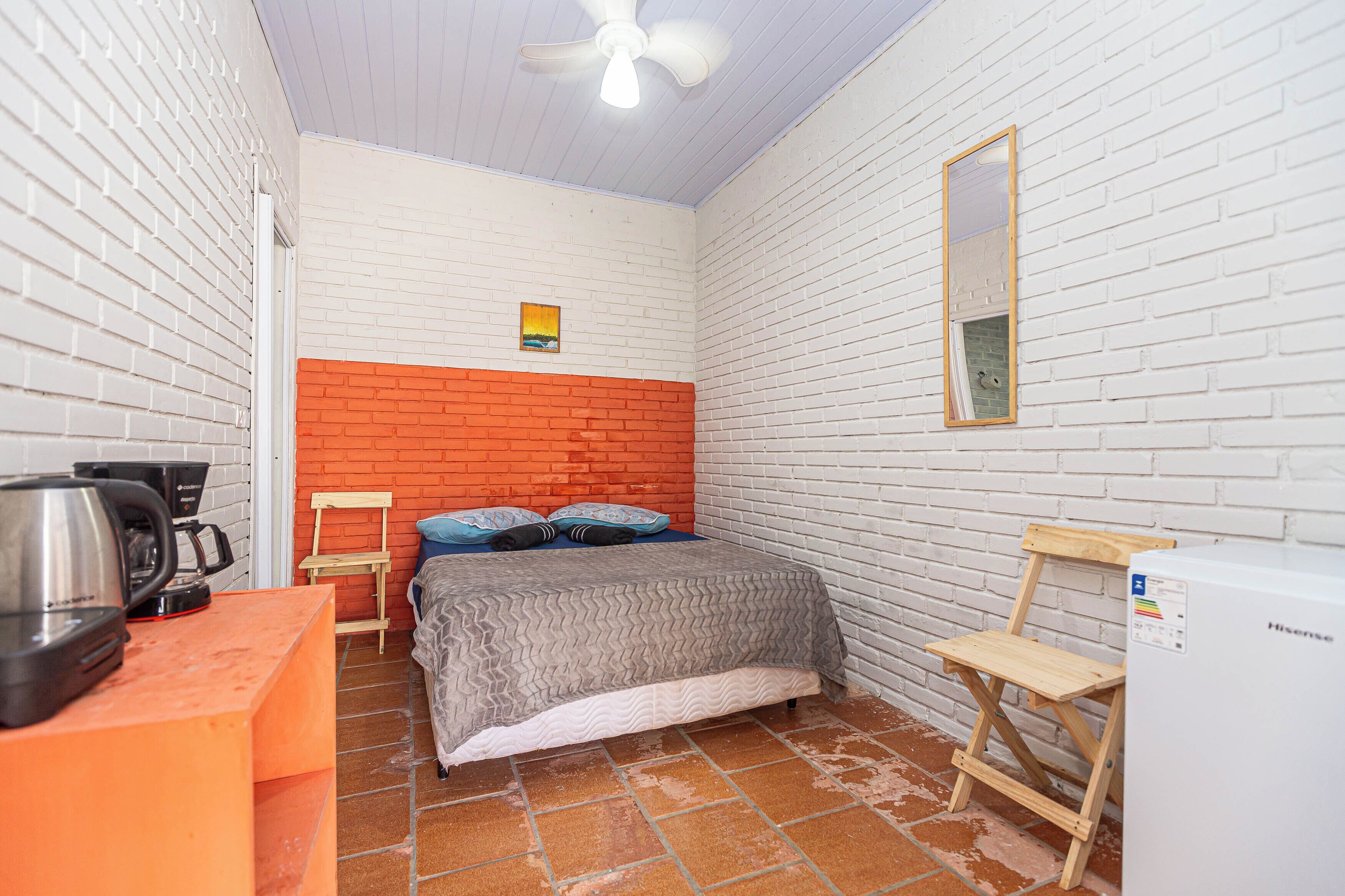 A compact room with a bed, a small table with a kettle, a chair, a wooden stool, and a white refrigerator.