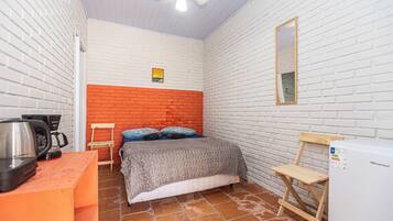 A compact room with a bed, a small table with a kettle, a chair, a wooden stool, and a white refrigerator.