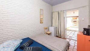 A bedroom with a white brick wall, a bed with a blue and grey blanket, a wooden chair, a small white fridge, and a wooden cabinet with a coffee maker.