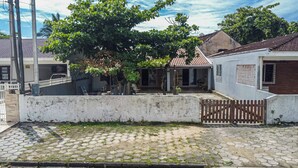 A residential area with a paved courtyard, a white wall with a gate, and a tree providing shade.