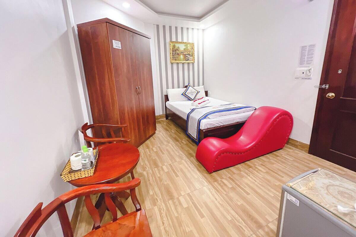 Een compacte kamer met een bed, een rode stoel, een houten garderobe, een klein tafeltje met toiletartikelen en een aan de muur hangend schilderij.