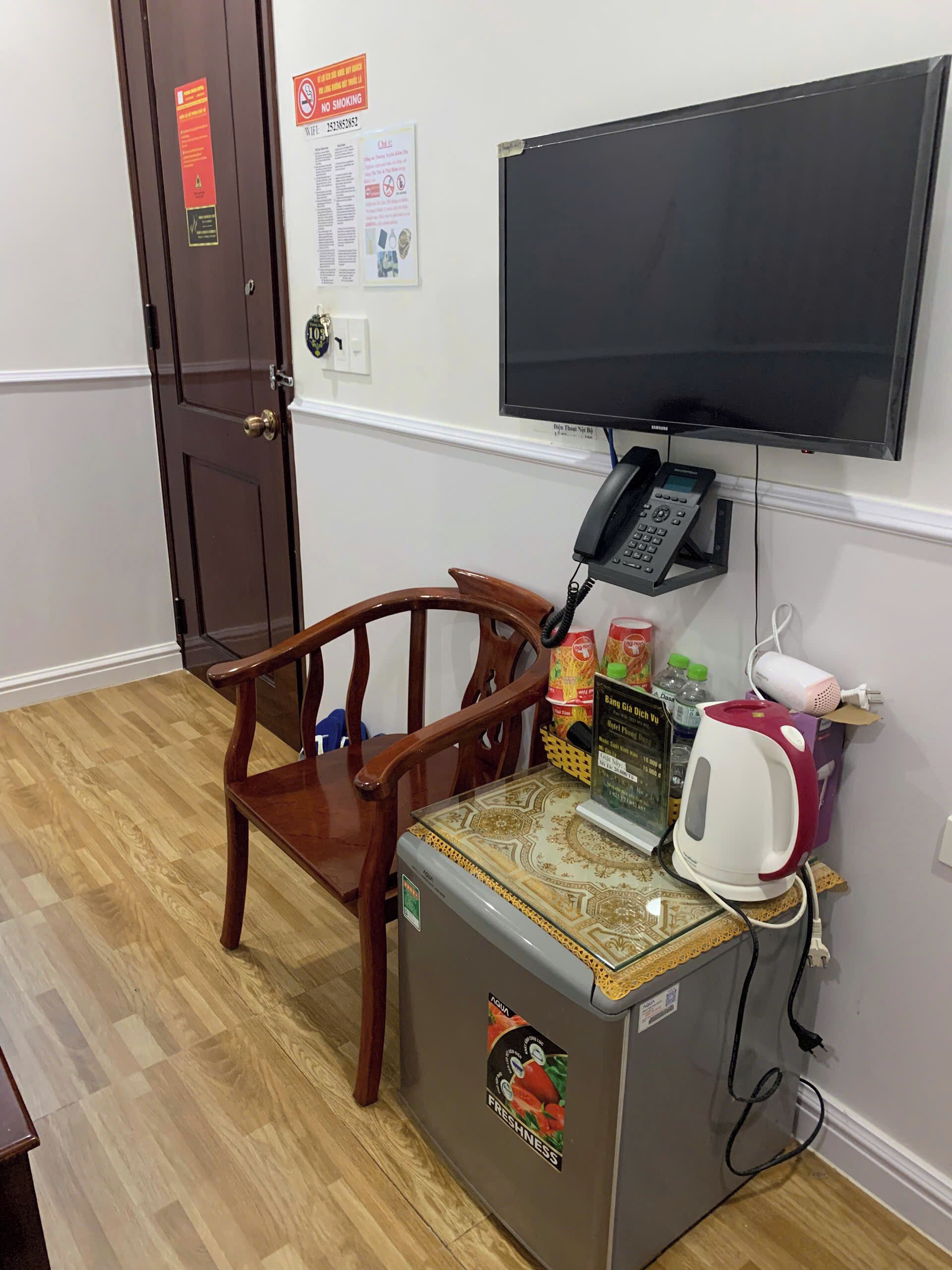A room with a wooden chair, a refrigerator, a microwave, a telephone, a television, and a wall-mounted power outlet.