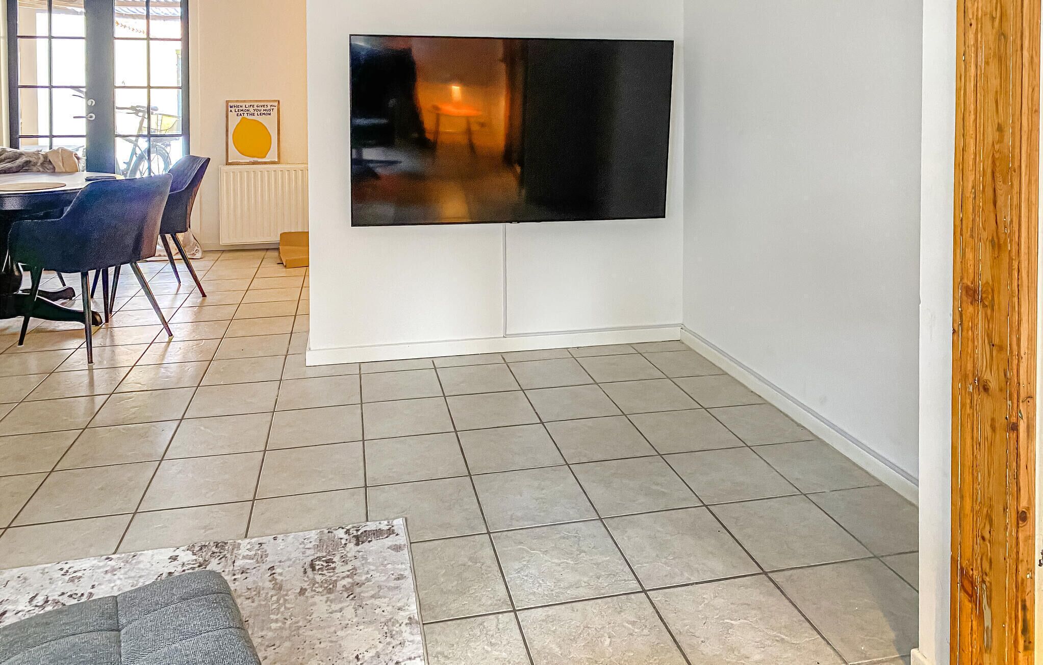 Una sala de estar moderna con un televisor de pantalla plana grande montado en la pared, un comedor con una mesa y sillas, y piso de baldosas.