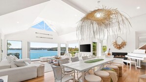 Un vaste espace de vie comprenant une grande table à manger, des canapés blancs et des poufs en osier. On aperçoit un piano au fond de la pièce, et de grandes fenêtres offrent une vue sur la mer.