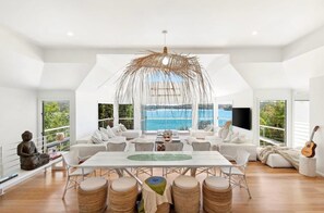 A spacious living area with a large table, white sofas, and a view of the sea. - Bundeena Sands Barefoot Private Beach Oasis (Bundeena)