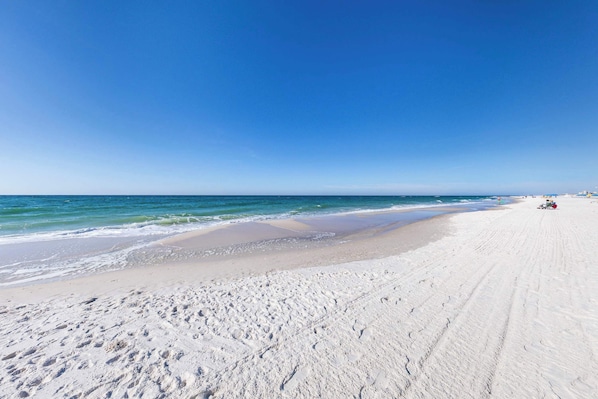 A wide sandy beach with gentle waves, a clear blue sky, and a distant cityscape. - *Heart of Orange Beach - Gulf front*Pool*Balcony (Orange Beach)