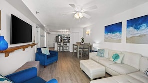 A modern living room with a flat-screen TV, blue and white furniture, a dining area with chairs and a table, and a kitchen in the background. - *Heart of Orange Beach - Gulf front*Pool*Balcony (Orange Beach)