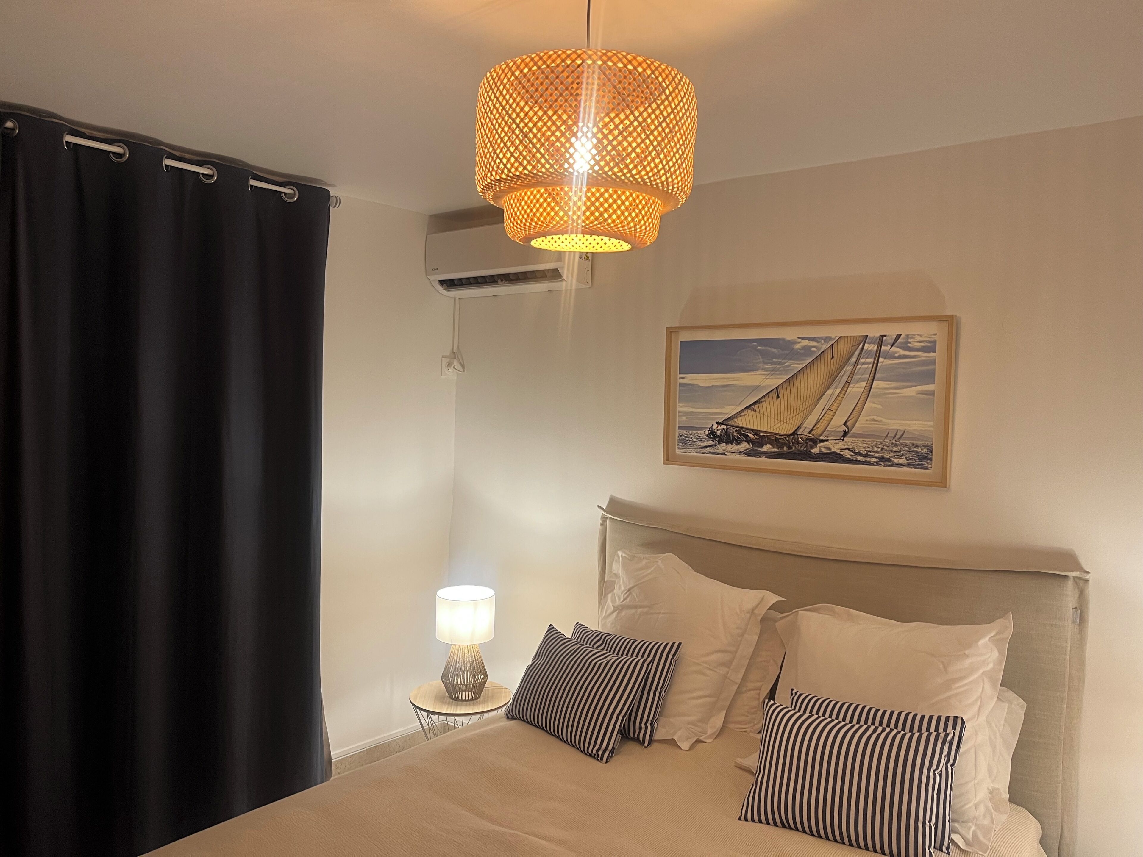 Een slaapkamer met een bed, een nachtkastje met een lamp, een schilderij van een zeilboot en een zwart gordijn.