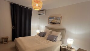 Quarto com cama, luminárias de cabeceira, luminária pendente, um pequeno banquinho e um banco de madeira.