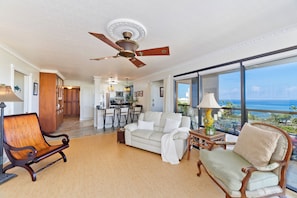 Amplia sala de estar con ventilador de techo alto, un ventanal con vista al océano y un rincón de descanso acogedor con un sofá y dos sillones.