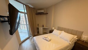A hotel room with a bed, a nightstand, a dresser, an air conditioning unit, and a view of buildings through a window with curtains. - Beach dream, sea and lagoon views, fully equipped apartment. (Rio de Janeiro)