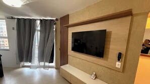 A modern living room with a large flat-screen TV mounted on a wooden wall, a built-in bench below, and a window with sheer curtains. - Beach dream, sea and lagoon views, fully equipped apartment. (Rio de Janeiro)