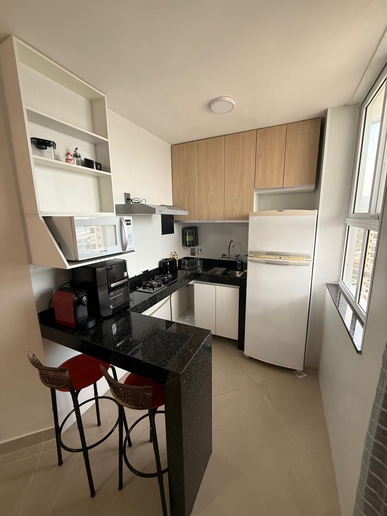 A compact kitchen with white cabinets, a black countertop, a built-in microwave, a stove, and a refrigerator. There are two red bar stools and a window providing natural light.