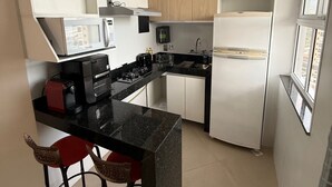 A compact kitchen with white cabinets, a black countertop, a built-in microwave, a stove, and a refrigerator. There are two red bar stools and a window providing natural light. - Beach dream, sea and lagoon views, fully equipped apartment. (Rio de Janeiro)