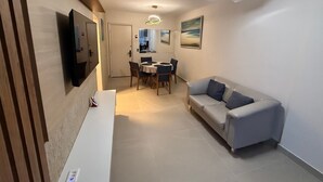 A modern living room with a sofa, a dining table with chairs, a TV mounted on the wall, and a ceiling fan. - Beach dream, sea and lagoon views, fully equipped apartment. (Rio de Janeiro)