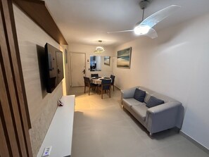 A modern living room with a sofa, a dining table with chairs, a TV mounted on the wall, and a ceiling fan. - Beach dream, sea and lagoon views, fully equipped apartment. (Rio de Janeiro)