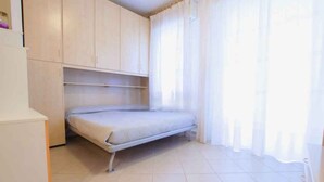 Een kleine, lichte kamer met een bed, een kledingkast en een raam met dunne gordijnen.