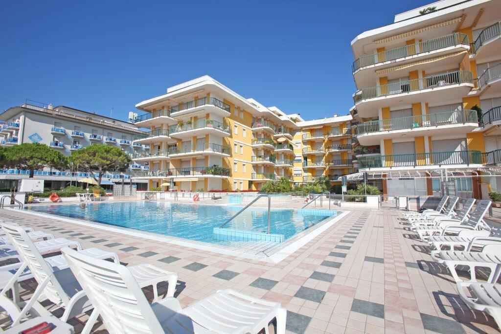 A swimming pool with lounge chairs, a building with multiple balconies, and a clear blue sky.
