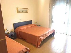 A bedroom with two beds, a nightstand with a lamp, a framed picture on the wall, and a window with curtains. - Holiday apartment mit Klimaanlage und Balkon (Lido di Jesolo)