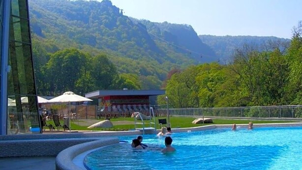 A swimming pool with people in it, surrounded by a building and a view of a forested hill. - Hotel Restaurant Piccolo (Thale)