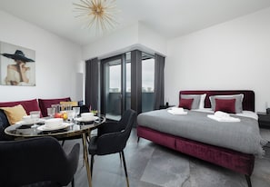 Moderni hotellihuone, jossa on suuri sänky, ruokapöytä tuoleineen, lasiovesta avautuva näkymä ja kaupunkimaisema, joka on nähtävissä ulkona.