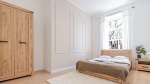 Un dormitorio con una cama de madera, un tapete blanco, un armario y una ventana con cortinas.