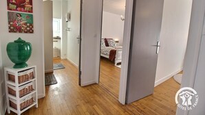 A hallway with wooden flooring leading to a bedroom with a bed and a nightstand. There is a green vase on a white shelf and a basket on the floor. - Les Hirondelles - LAURENAN (LAURENAN)