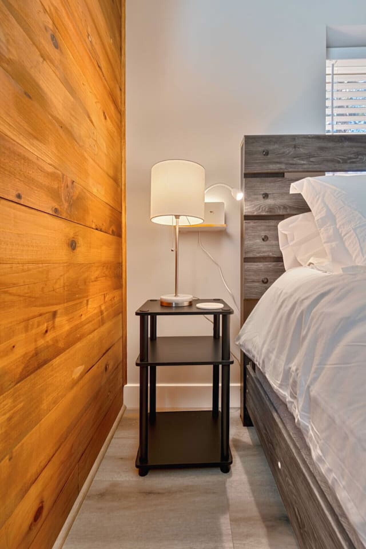 Un dormitorio con pared de madera, una mesita de noche con lámpara y una cama con ropa blanca de cama.