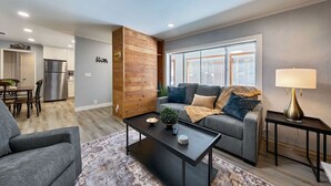Ein modernes Wohnzimmer mit grauem Sofa, Couchtisch und Lampe. Im Hintergrund befindet sich ein Küchenbereich mit Kühlschrank und Schränken.