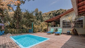 A wooden deck with a swimming pool, lounge chairs, and a covered area with a view of trees. - Casa em Condom nio com Piscina e rea Gourmet (Garopaba)