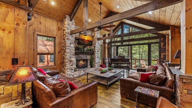 A cozy cabin interior with a stone fireplace, leather sofas, and a large window overlooking the forest. - Big Bear Falls - Riverfront | Pet Friendly | Hot Tub (Ellijay)