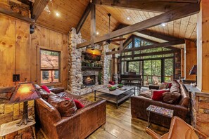 L’intérieur chaleureux d’un chalet avec une cheminée en pierre, des canapés en cuir et une grande fenêtre donnant sur la forêt.