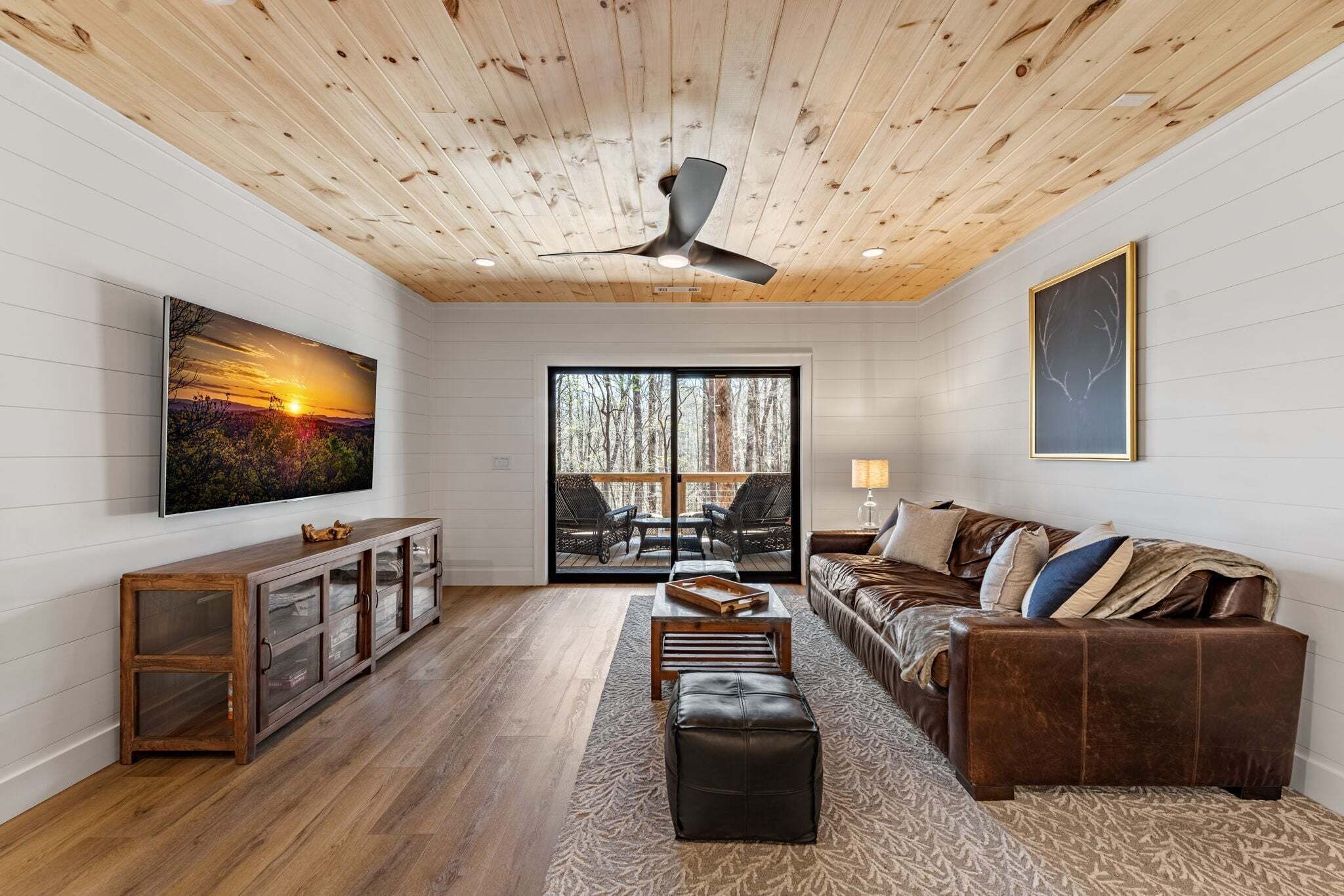 A living room with a wooden ceiling, a large TV, a sofa, a coffee table, and a view of trees through a sliding door.