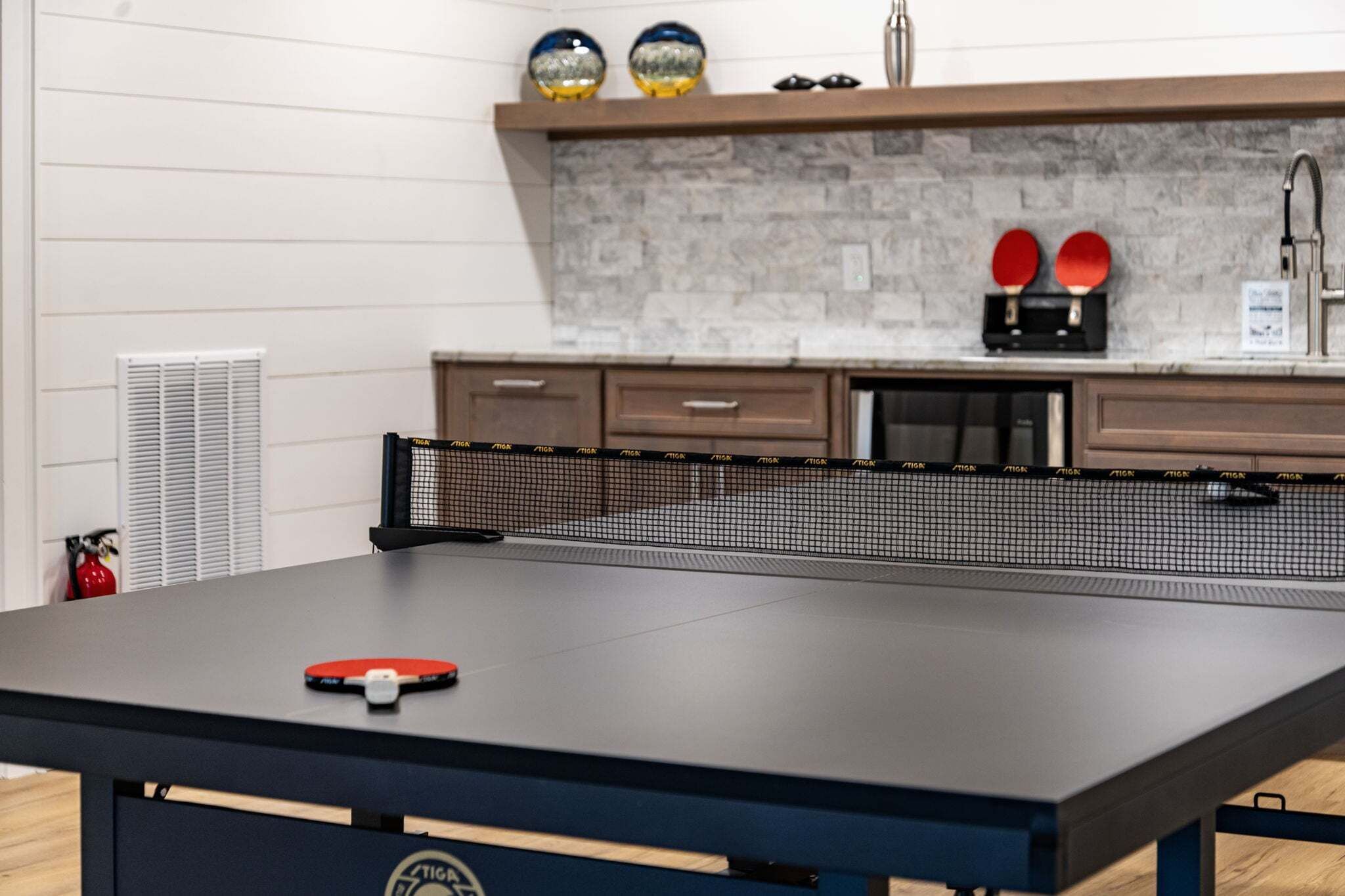 A ping pong table with a net, a red paddle, and two red paddles on a shelf above.