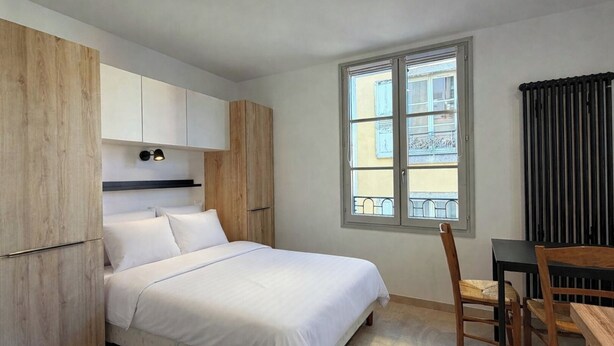 Ein Schlafzimmer mit Bett, Schreibtisch und Stuhl. Ein Fenster bietet Blick auf Gebäude.