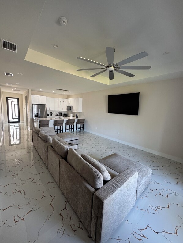Un salon moderne avec un grand canapé d’angle, un ventilateur de plafond et une cuisine en arrière-plan.