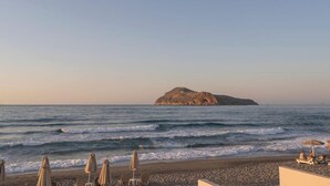 A beach with lounge chairs and umbrellas, a rocky island in the distance, and calm waves. - Marika Hotel Chania (Platanias)