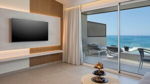 A modern hotel room with a large flat-screen TV mounted on the wall, a balcony with a view of the sea, and a round coffee table with pastries. - Marika Hotel Chania (Platanias)