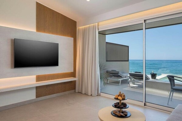 Habitación de hotel moderna con un televisor de pantalla plana grande montado en la pared, un balcón con vistas al mar y una mesa de centro redonda con pasteles.
