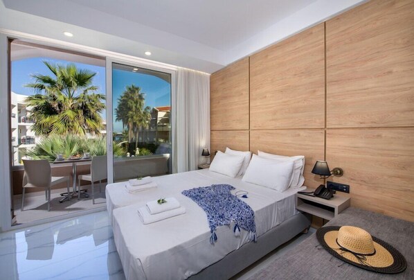 A modern hotel room with a large bed, a desk with chairs, a balcony with palm trees, and a view of other buildings. - Marika Hotel Chania (Platanias)