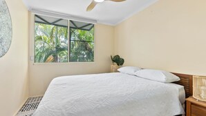 Una camera da letto con un letto, una finestra vista palme, una ventola a soffitto e un comodino con una lampada.